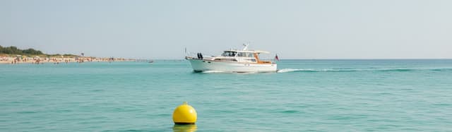 un bateau de plaisance à moteur naviguant lentement près d'un rivage l'été, plage et baigneurs au loin, bouée jaune sphé