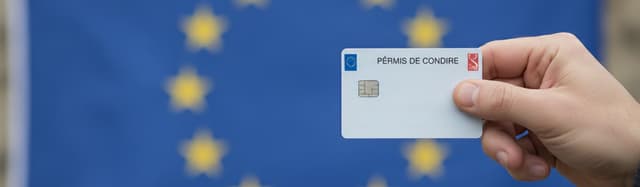 main tendant un permis de conduire français format carte de crédit, drapeau européen flouté en arrière-plan