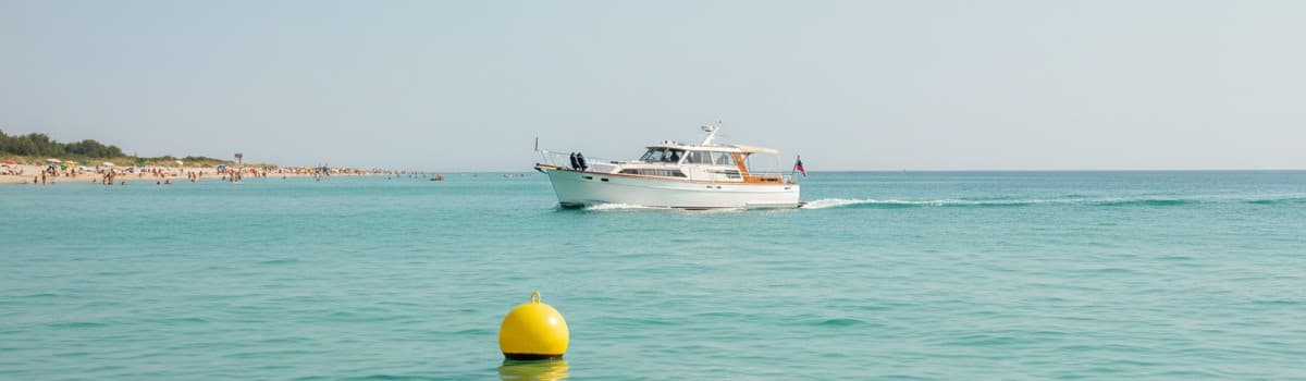 un bateau de plaisance à moteur naviguant lentement près d'un rivage l'été, plage et baigneurs au loin, bouée jaune sphé