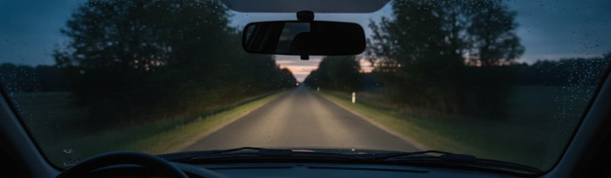 vue depuis l'intérieur d'une voiture au crépuscule, pare-brise occupant tout le cadre, route départementale bordée d'arb