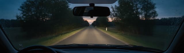 vue depuis l'intérieur d'une voiture au crépuscule, pare-brise occupant tout le cadre, route départementale bordée d'arb