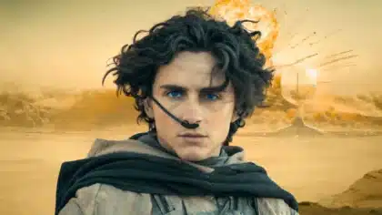 Un jeune homme aux yeux bleus intenses et cheveux bouclés noirs porte une tenue désertique sombre avec un stillsuit, sur fond de dunes dorées et d'explosion orangée.